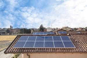 Solar panels installed on the tiled roofs of residential buildings in St. John the Baptist Parish under a partly cloudy sky, with a town and open field in the background.